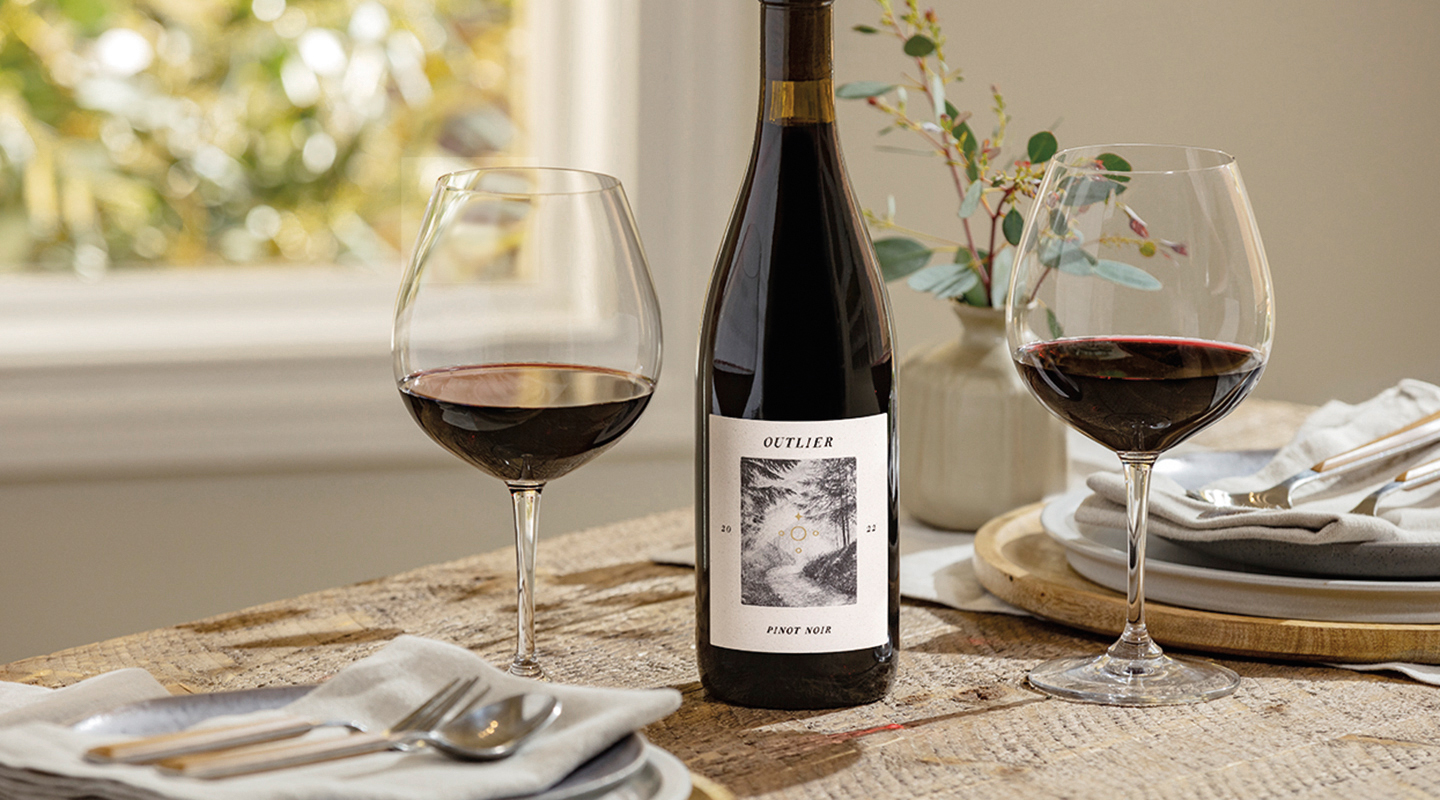 Outlier Pinot Noir bottle on table with two filled wineglasses and table settings with window view in background